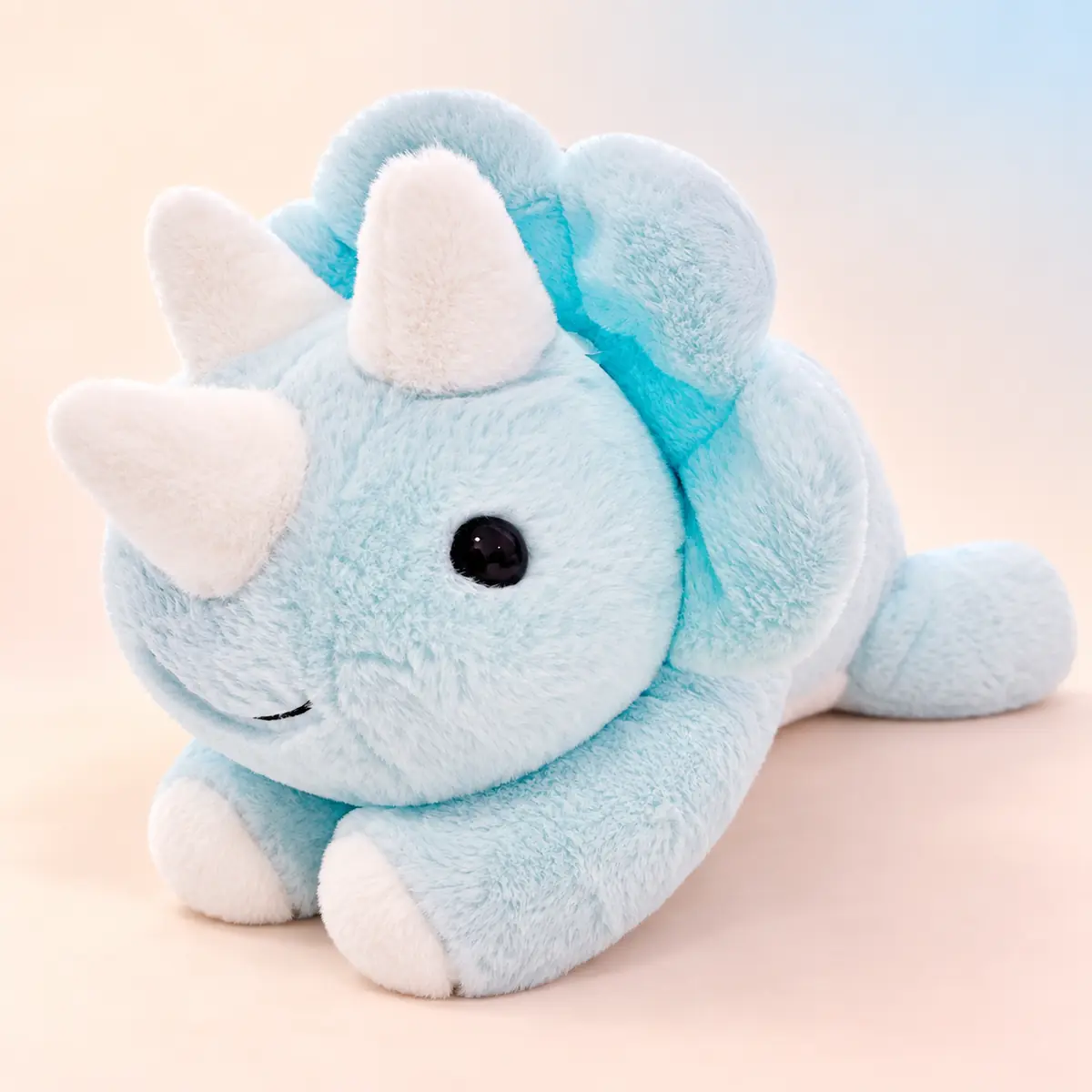 Rex, the pale blue triceratops dinosaur weighted plush, lying down with soft white horns against a soft peach studio background